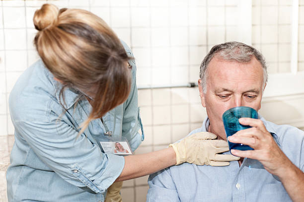 ** ID card is completely created by the photographer. Focus on nurse's hand and throat ** Healthcare worker. wearing surgical gloves, checking man's throat as he drinks. She is a Speech Therapist specialising in dysphagia (swallowing) and she is checking the man's swallow as he drinks. She is looking for laryngial extrusian and the movement of the Adam's apple. Neutral tiled background could be in a hospital or could be a home visitMore like this